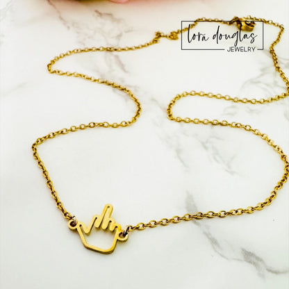A Middle Finger Necklace by Lora Douglas, crafted from polished stainless steel, is displayed on a white marble surface. The Lora Douglas logo appears in the top right corner.