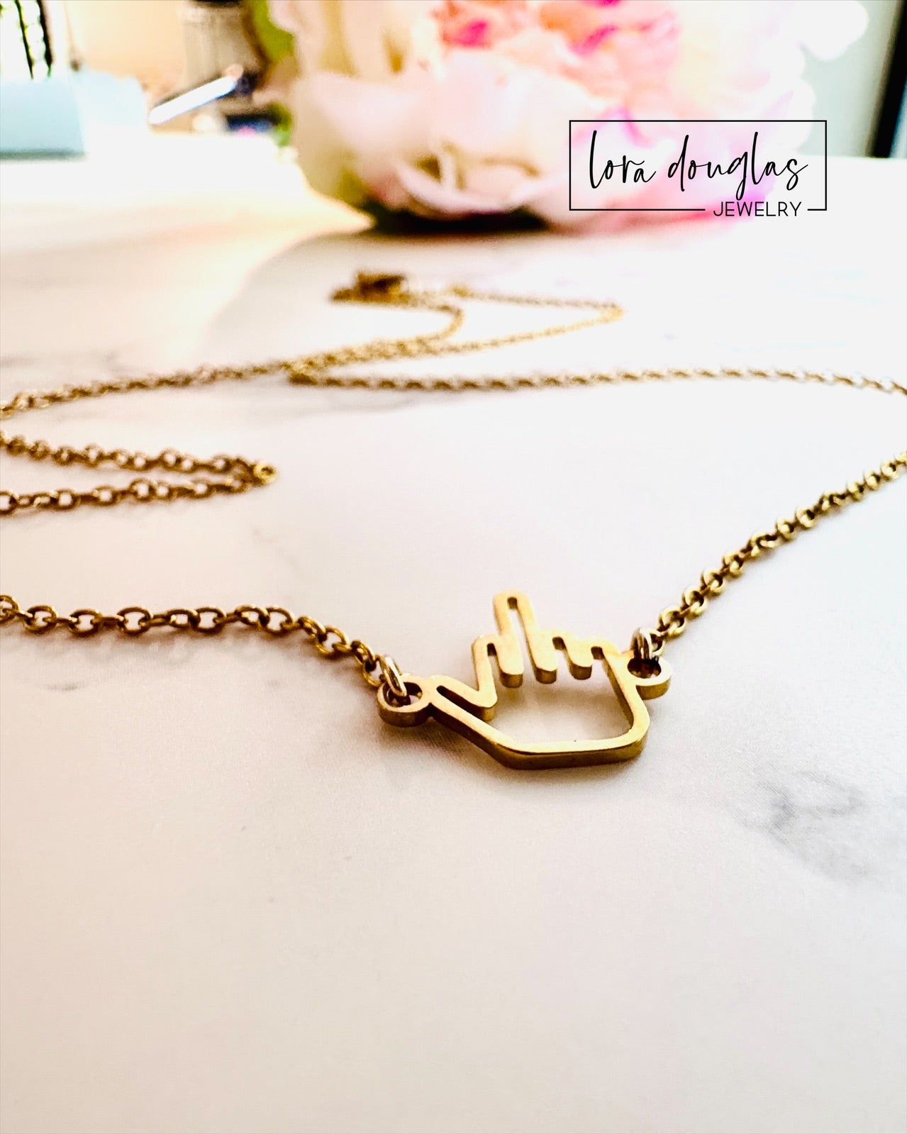 The Middle Finger Necklace by Lora Douglas features a playful gold middle finger charm, shown on a light surface with a blurred flower in the background. Lora Douglas Jewelry appears in the image text.