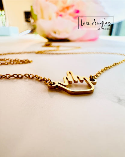 The Middle Finger Necklace by Lora Douglas features a polished stainless steel middle finger charm on a gold chain, presented on a white background with a soft pink flower and the Lora Douglas logo above.
