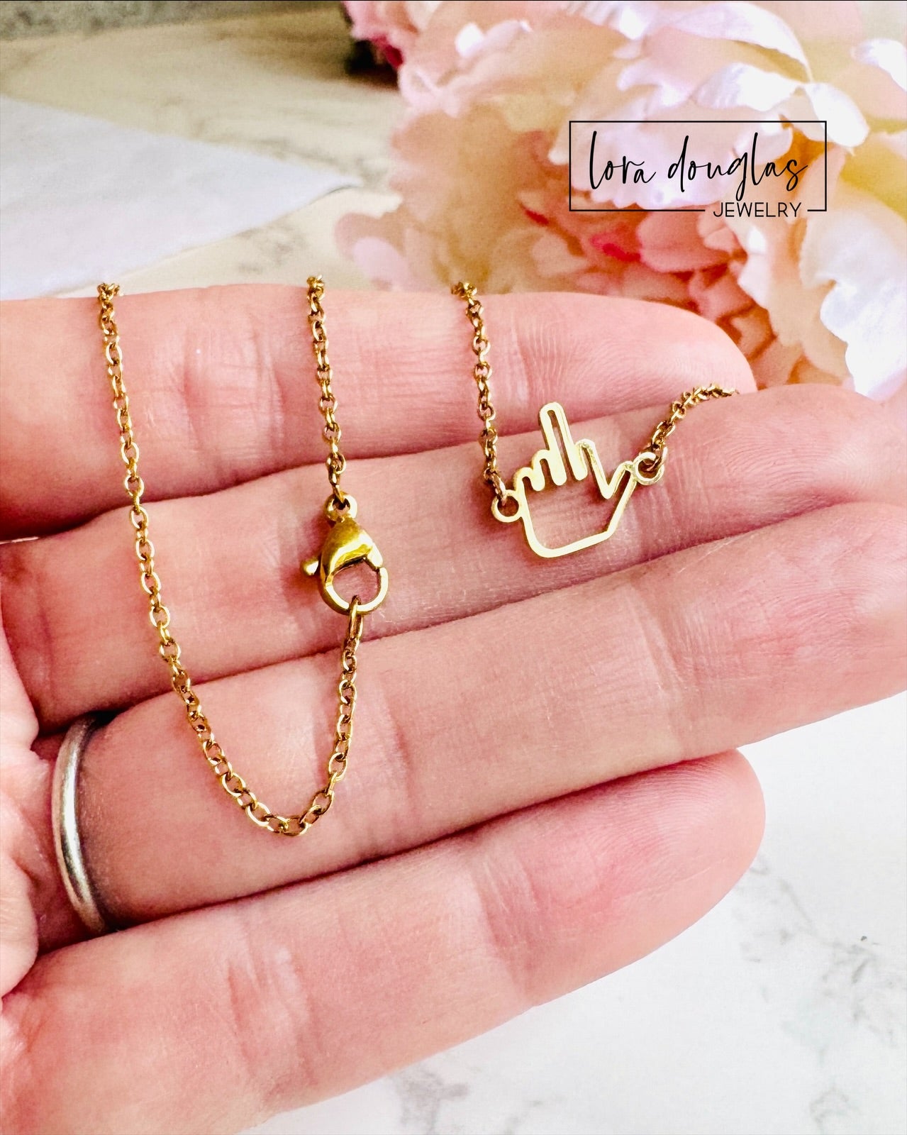A hand holds the Lora Douglas Middle Finger Necklace with a playful gold middle finger charm. A soft pink flower is in the background, and the Lora Douglas logo is in the top right corner.