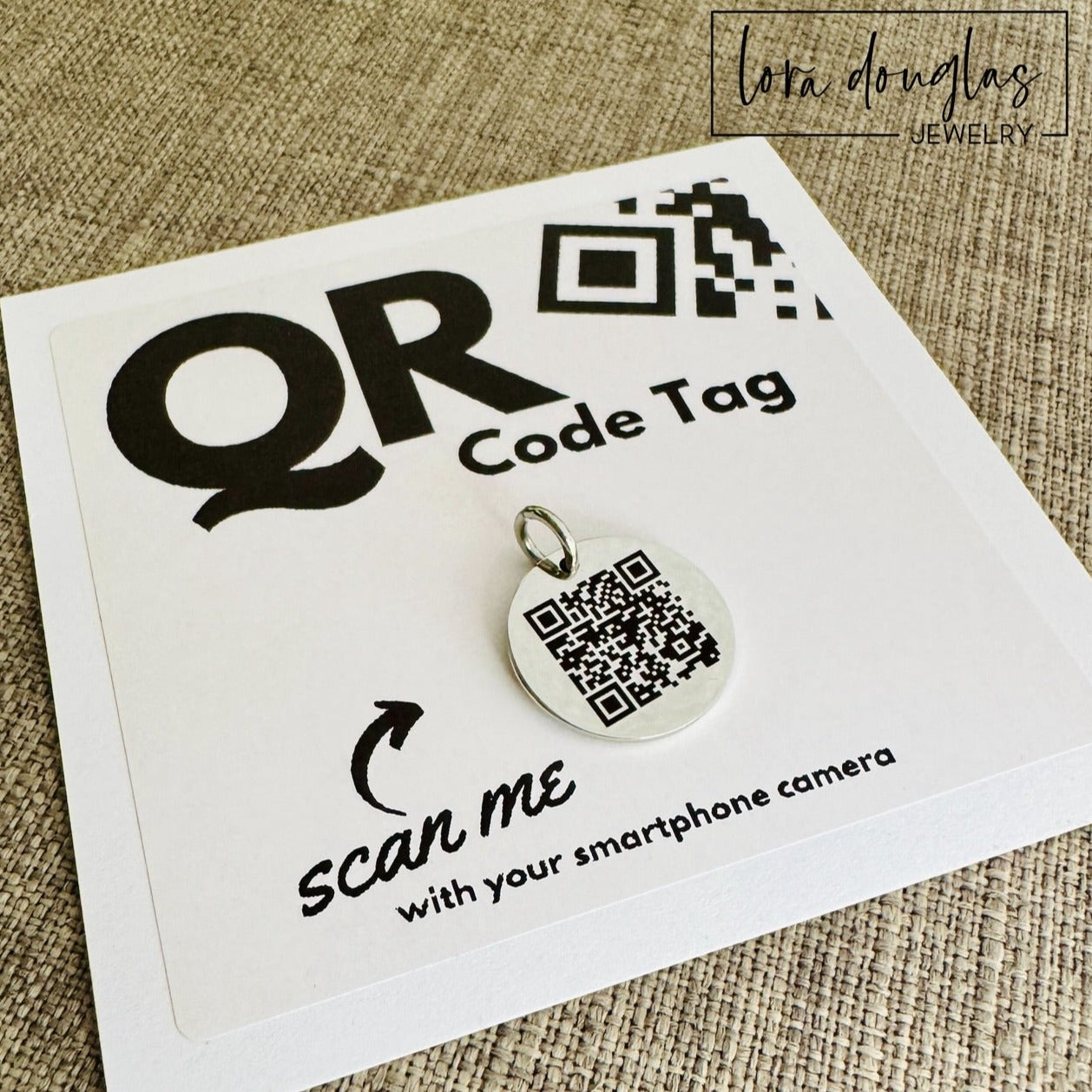 A stainless steel QR code tag with a lobster clasp, displayed on a white background with a smartphone in the background, indicating how to scan the code.