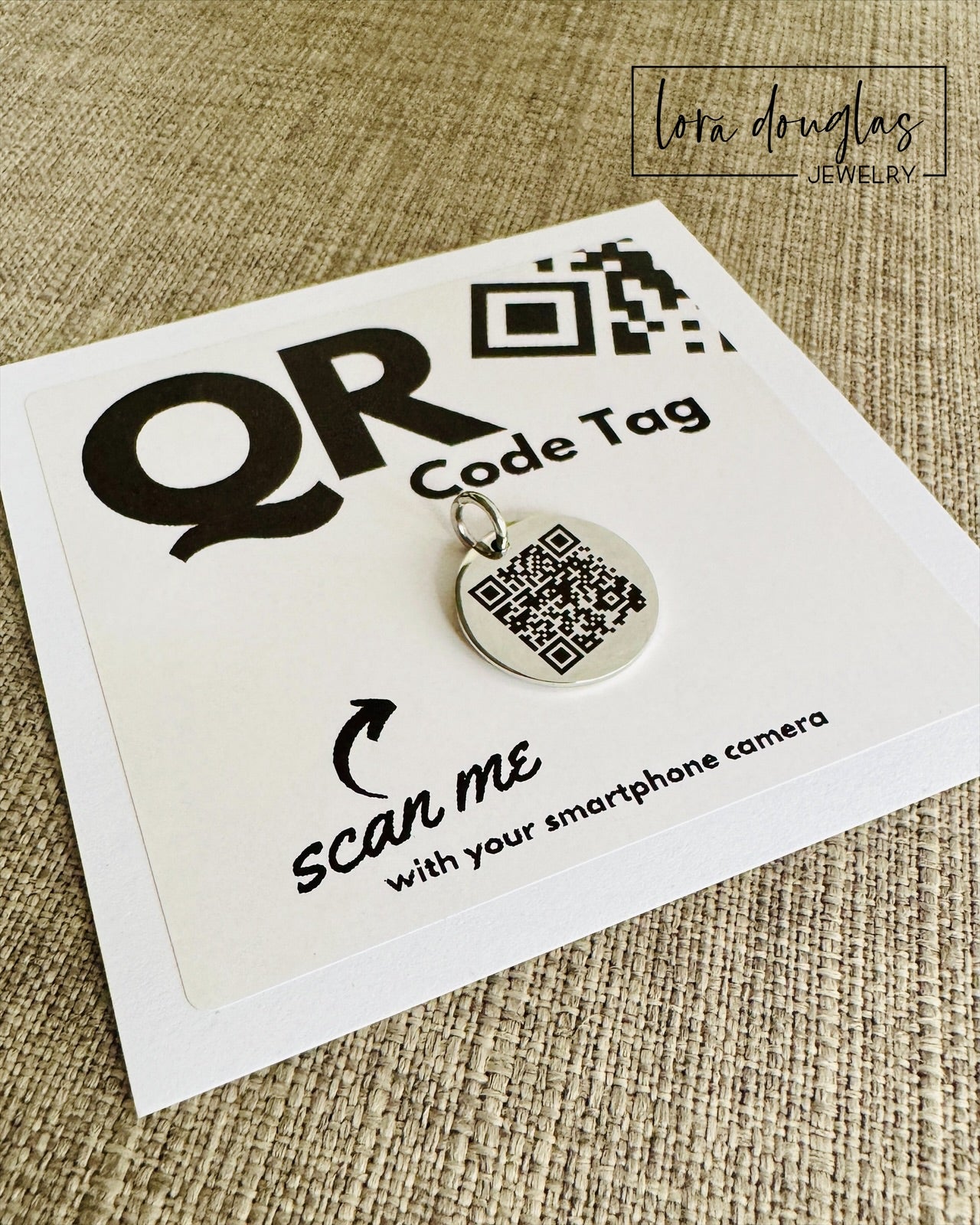 A stainless steel QR code tag with a lobster clasp, displayed on a white background with a smartphone in the background, indicating how to scan the code.