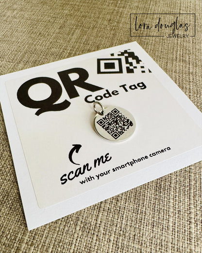 A stainless steel QR code tag with a lobster clasp, displayed on a white background with a smartphone in the background, indicating how to scan the code.