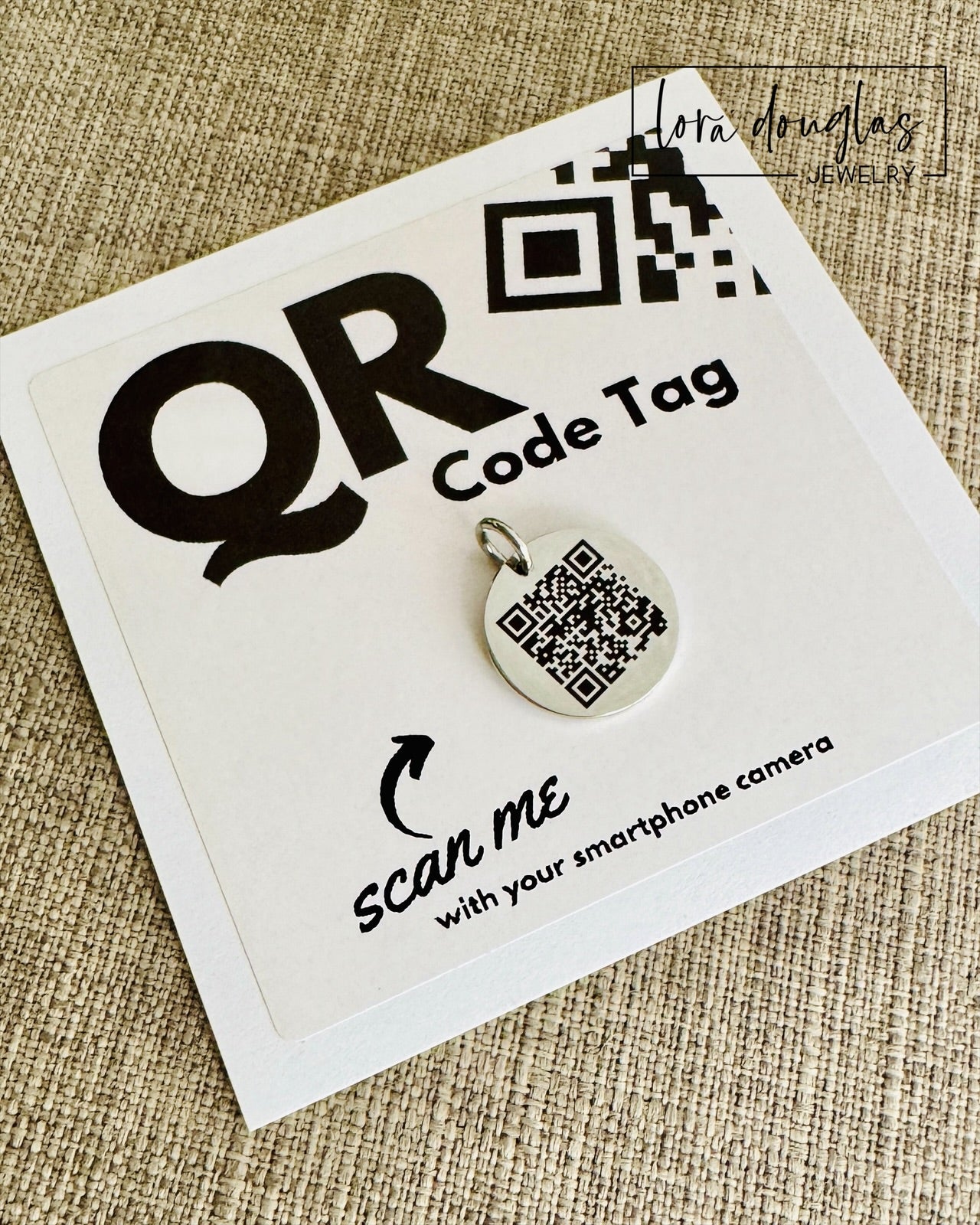 A stainless steel QR code tag with a lobster clasp, displayed on a white background with a smartphone in the background, indicating how to scan the code.