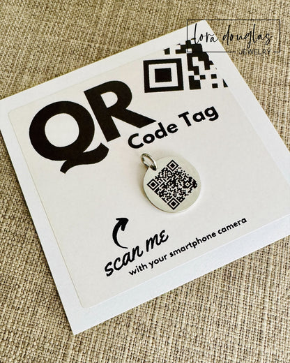 A stainless steel QR code tag with a lobster clasp, displayed on a white background with a smartphone in the background, indicating how to scan the code.