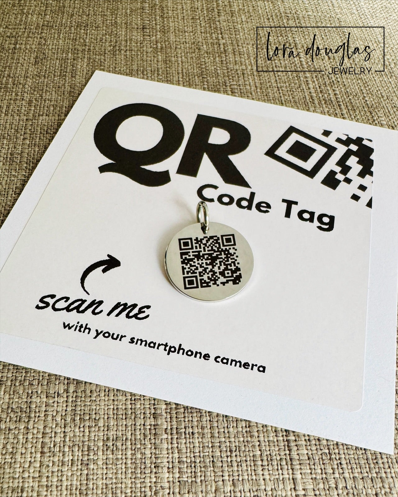 A stainless steel QR code tag with a lobster clasp, displayed on a white background with a smartphone in the background, indicating how to scan the code.