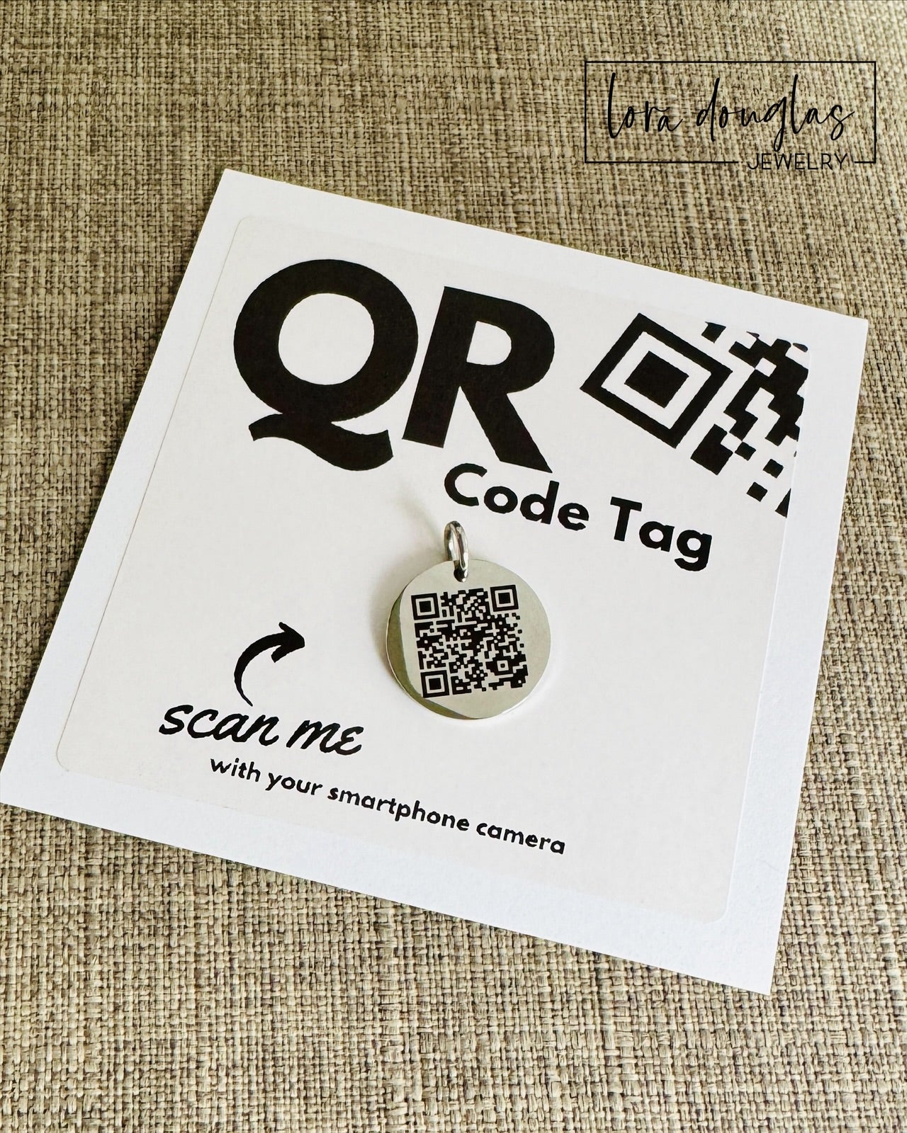 A stainless steel QR code tag with a lobster clasp, displayed on a white background with a smartphone in the background, indicating how to scan the code.