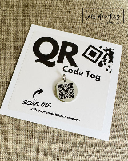 A stainless steel QR code tag with a lobster clasp, displayed on a white background with a smartphone in the background, indicating how to scan the code.