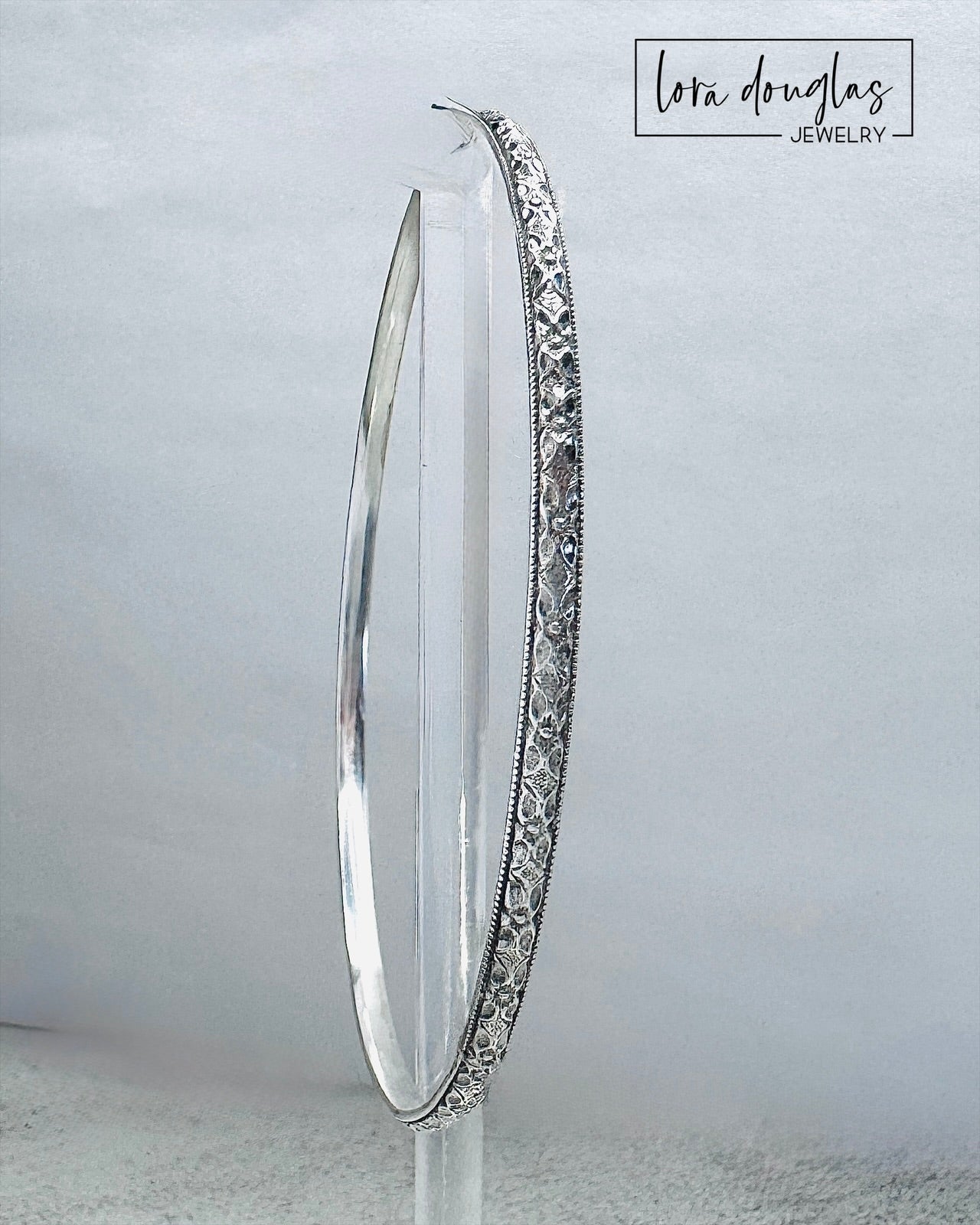 Handcrafted sterling silver bangle bracelet with textured and polished finish displayed on clear stand.