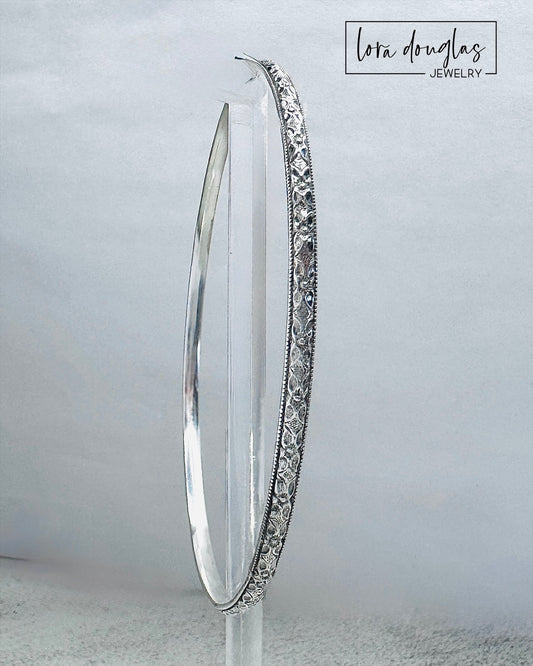 Handcrafted sterling silver bangle bracelet with textured and polished finish displayed on clear stand.