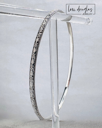 Handcrafted sterling silver bangle bracelet with textured and polished finish displayed on clear stand.