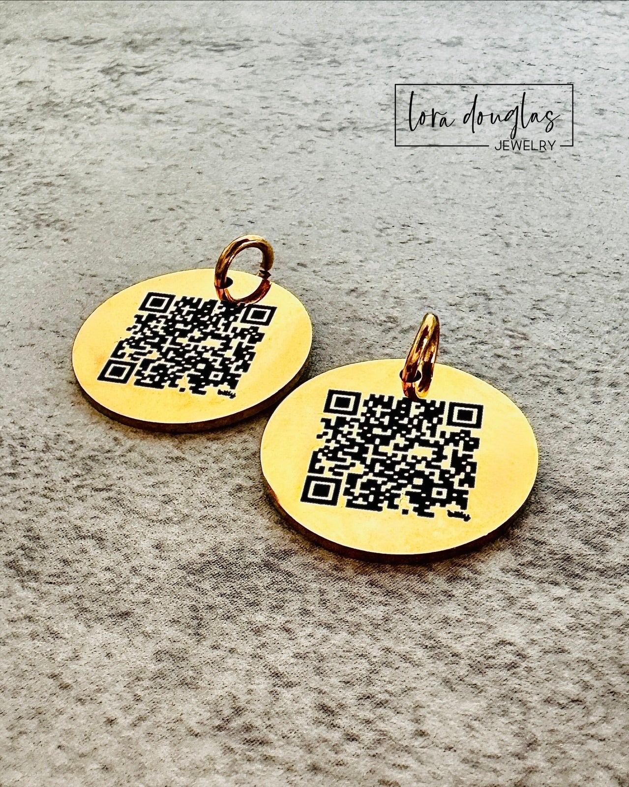 Gold charm with QR code on a gray surface, featuring 'Lora Douglas Jewelry' branding.