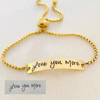 A gold bracelet with a bar pendant engraved with the handwritten message 'Love you more'.