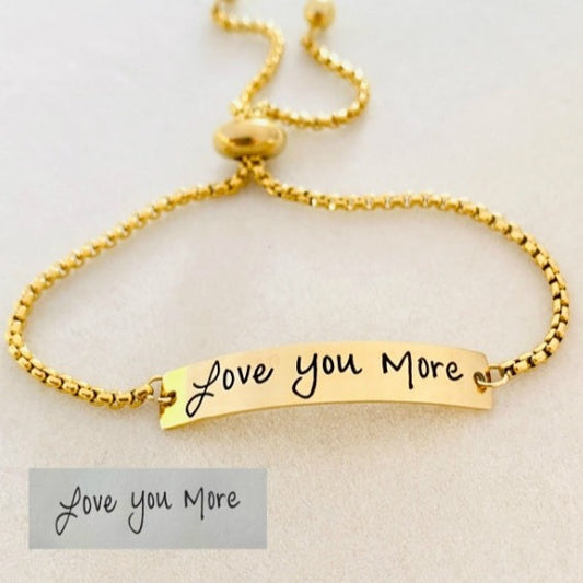 A gold bracelet with a bar pendant engraved with the handwritten message 'Love you more'.
