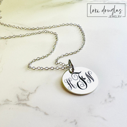 A silver monogram necklace with an engraved initial pendant laid out on a marble surface.