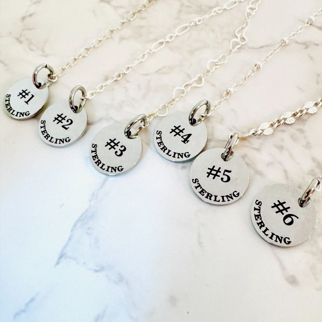 Six “Permanent Jewelry ID Tags for Displays” by Lora Douglas Jewelry, labeled “#1 STERLING” to “#6 STERLING,” are arranged in a row on white marble (chains not included).