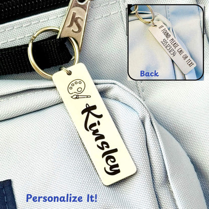 A personalized stainless steel ID tag with the name 'Kinsley' engraved on it, attached to a keychain and displayed on a backpack.