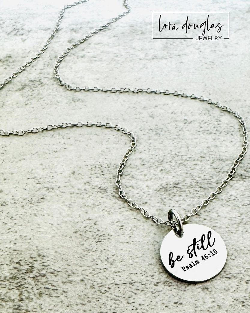 Silver necklace with a round pendant featuring 'be still' and Psalm 46:10 on a textured surface.