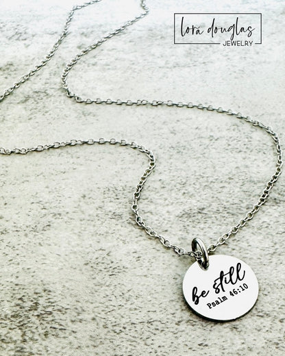 Silver necklace with a round pendant featuring 'be still' and Psalm 46:10 on a textured surface.