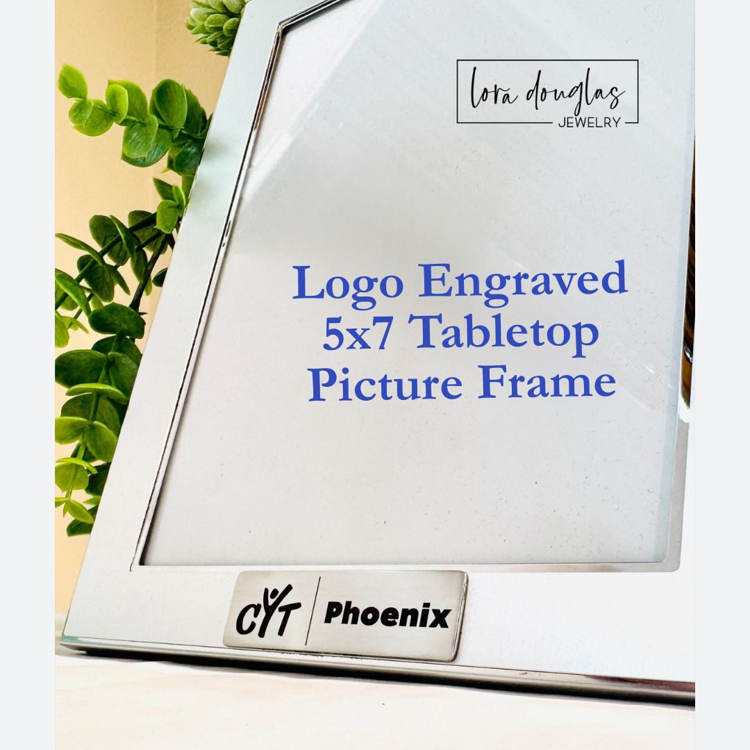A silver 5x7 tabletop picture frame with a logo engraved on the front, displayed on a table with a plant in the background.