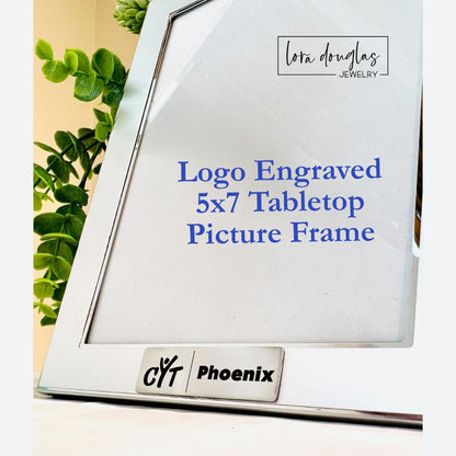 A silver 5x7 tabletop picture frame with a logo engraved on the front, displayed on a table with a plant in the background.
