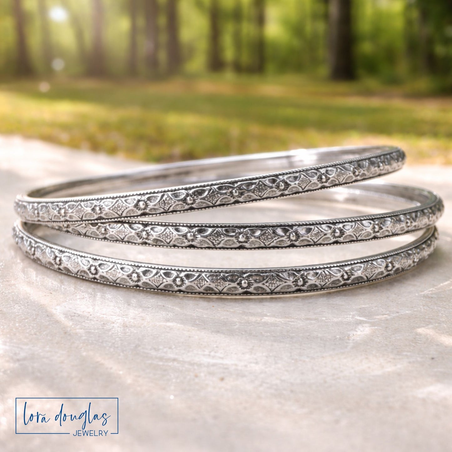Three sterling silver bangles stacked on a stone surface with a blurred natural background