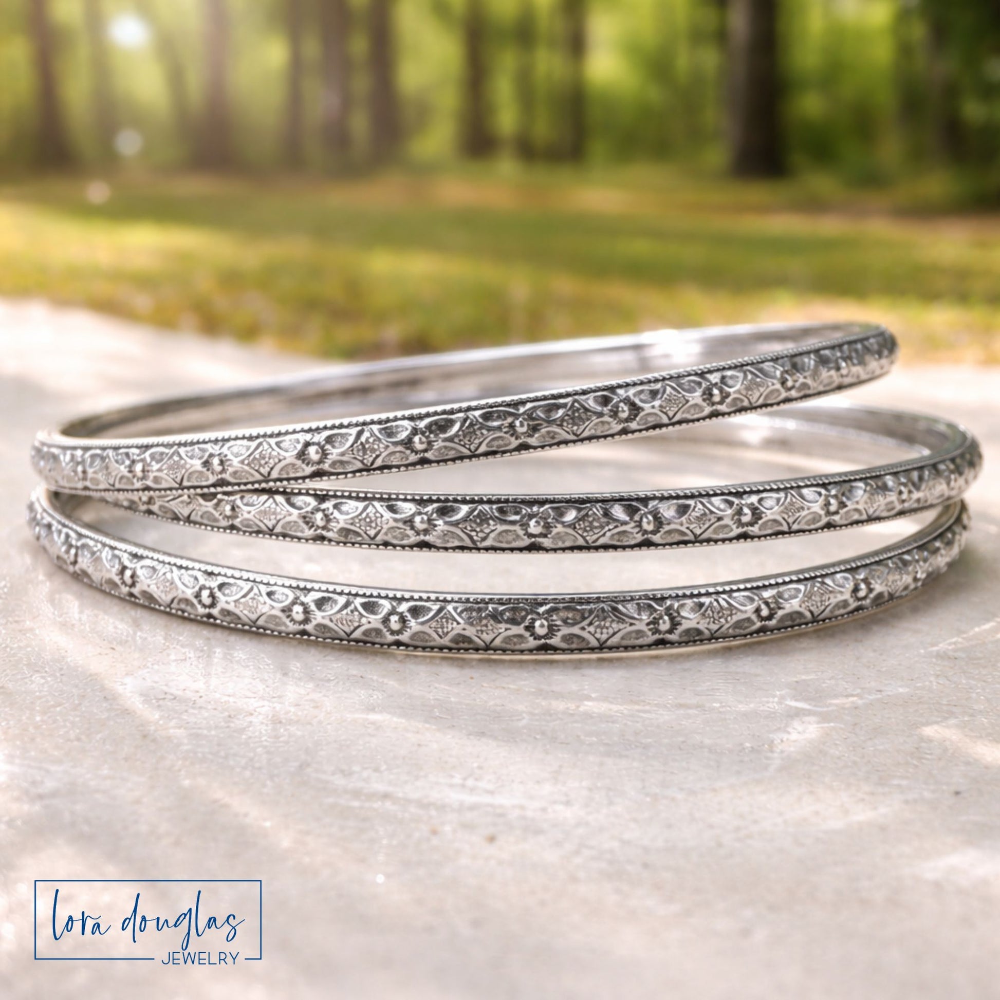 Three sterling silver bangles stacked on a stone surface with a blurred natural background