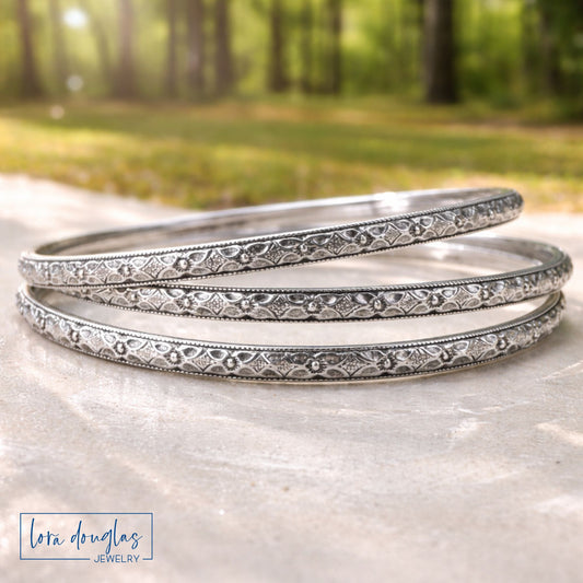 Three sterling silver bangles stacked on a stone surface with a blurred natural background