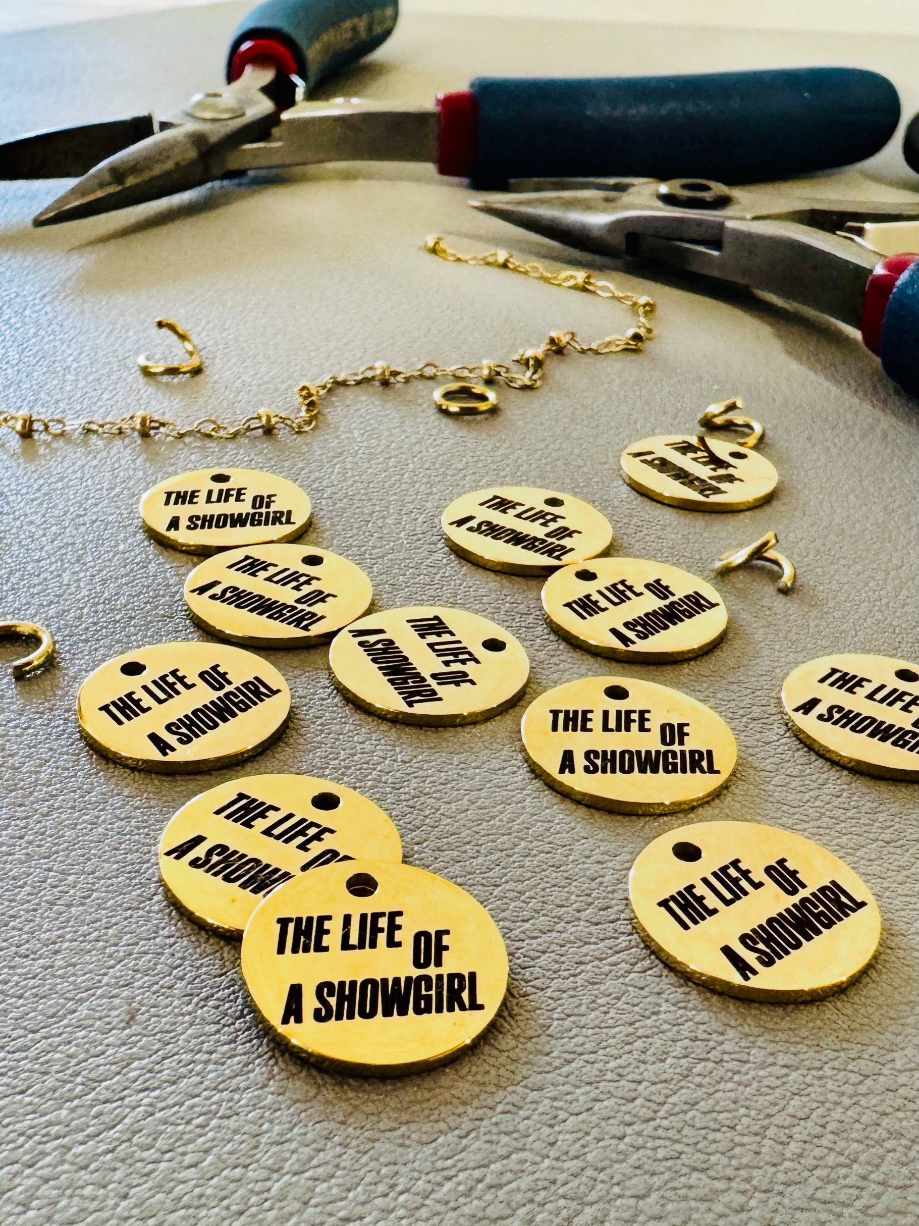 Gold tags with 'The Life of a Showgirl' text on a gray surface with pliers.