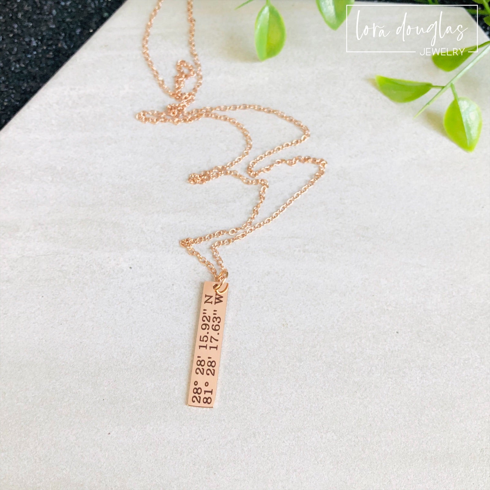 A rose gold necklace with a personalized engraving of a latitude and longitude on a pendant, displayed in a box with a blue and white design on the interior lining.