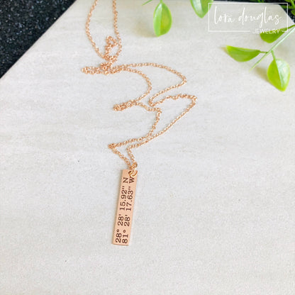 A rose gold necklace with a personalized engraving of a latitude and longitude on a pendant, displayed in a box with a blue and white design on the interior lining.