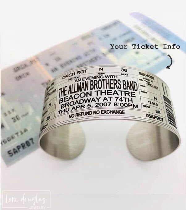 Close view of a personalized concert ticket bracelet showing the custom engraved artist, tour, venue and event information with a decorative barcode and ticket style details on a wide stainless steel cuff.
