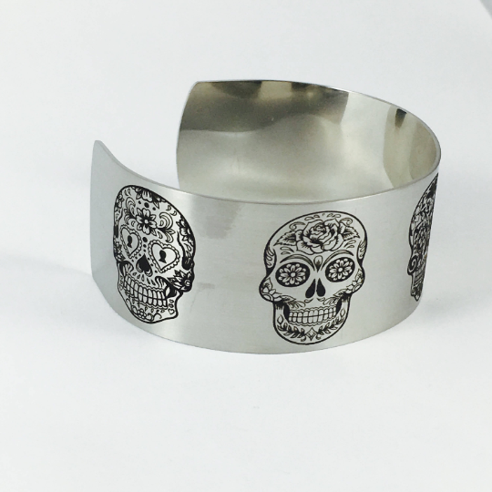 Close view of engraved sugar skull artwork on a wide stainless steel cuff bracelet, showing the detailed permanent engraving and matte finish of this distinctive skull jewelry design available in silver or gold.
