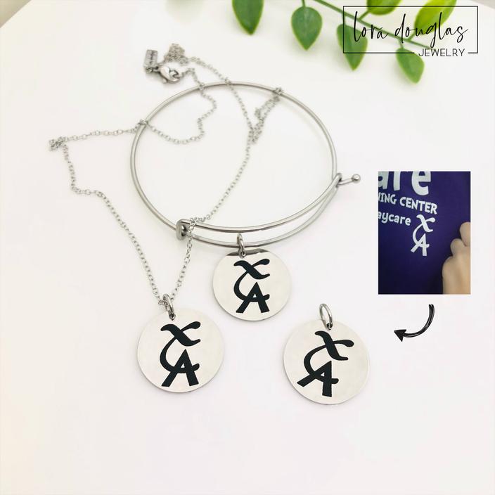 Custom drawing jewelry shown as an engraved round charm with clean black artwork against polished stainless steel, available as a charm only, 18 inch necklace, or bangle bracelet in silver, gold, and rose gold finishes