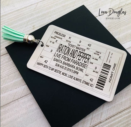 A stainless steel bookmark with laser engraved personalized text, depicting a concert ticket design, placed on a surface with a black bookmark and a green leaf in the background.