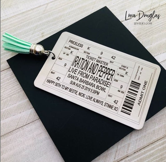 A stainless steel bookmark with laser engraved personalized text, depicting a concert ticket design, placed on a surface with a black bookmark and a green leaf in the background.