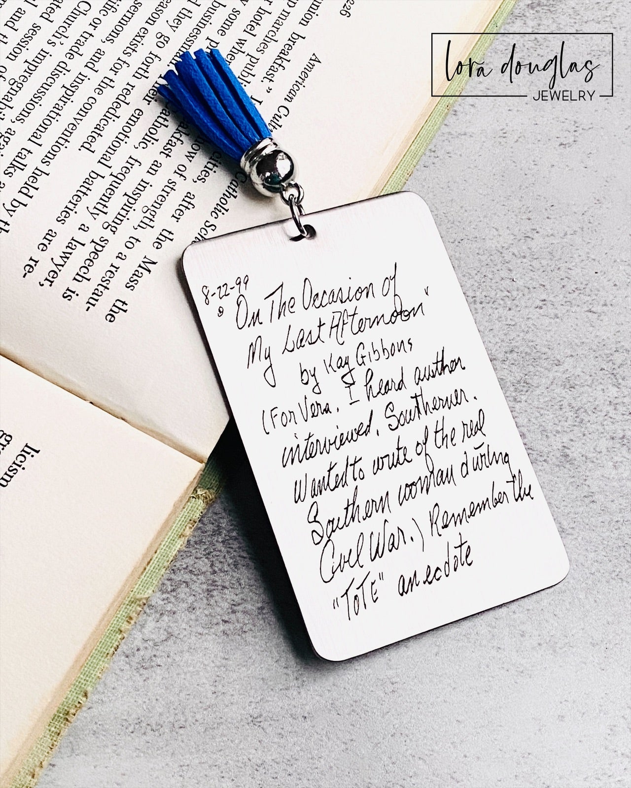A custom engraved bookmark with a handwritten message engraved on the metal. The bookmark is silver with black engraving and has a navy blue Tassel.