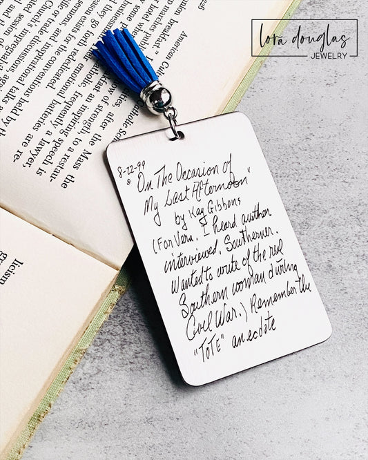 A custom engraved bookmark with a handwritten message engraved on the metal. The bookmark is silver with black engraving and has a navy blue Tassel.