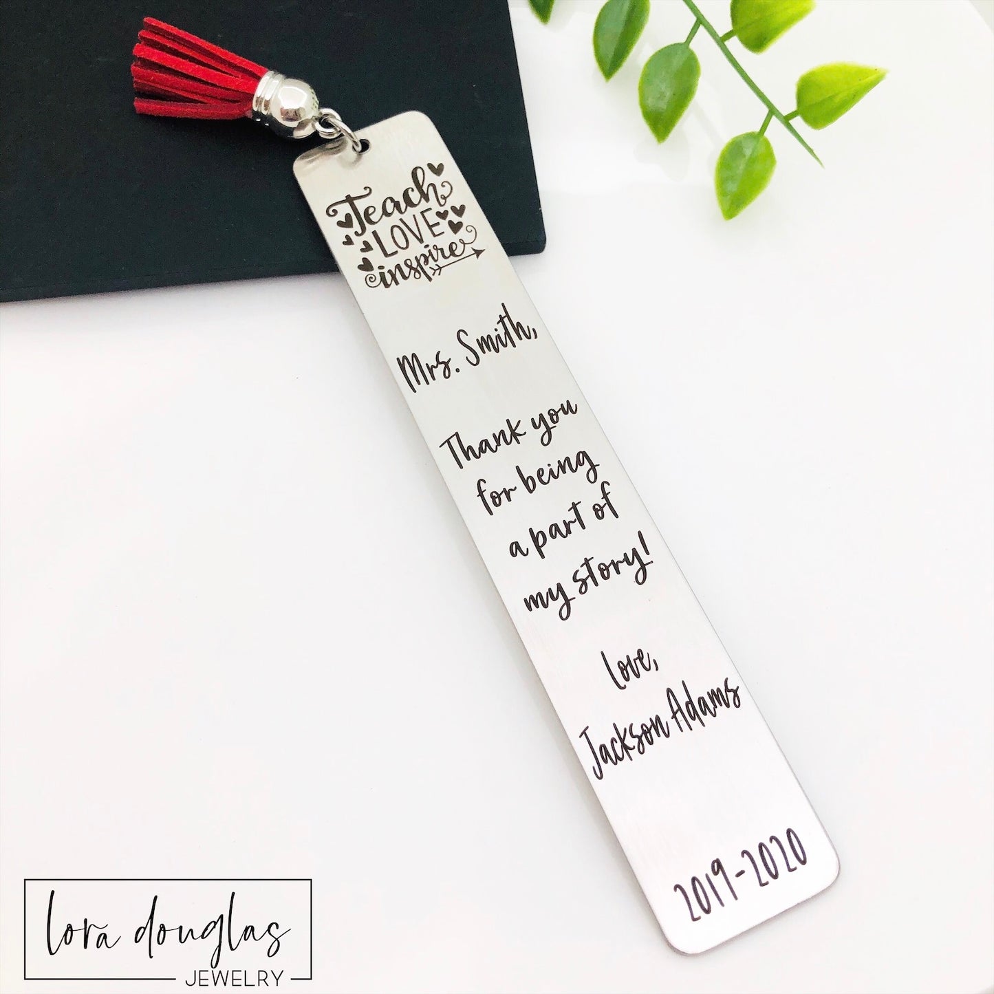 Engraved stainless steel teacher bookmark inside book with tassel visible  stainless steel bookmarks with the phrases 'Teach Love Inspire' and 'Thank you for being my teacher' engraved on them, presented in a navy blue velvet pouch with tassels.
