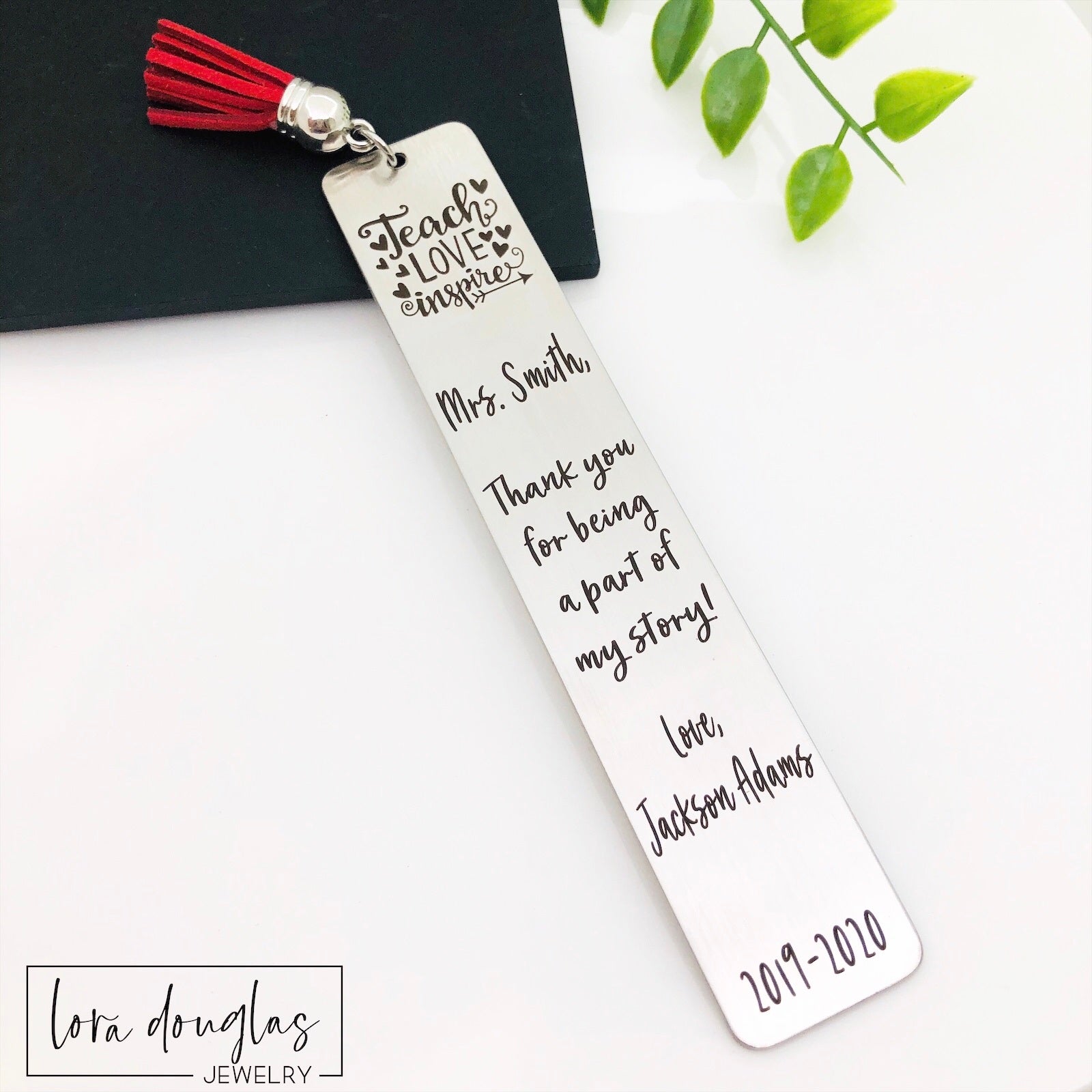 Engraved stainless steel teacher bookmark inside book with tassel visible  stainless steel bookmarks with the phrases 'Teach Love Inspire' and 'Thank you for being my teacher' engraved on them, presented in a navy blue velvet pouch with tassels.
