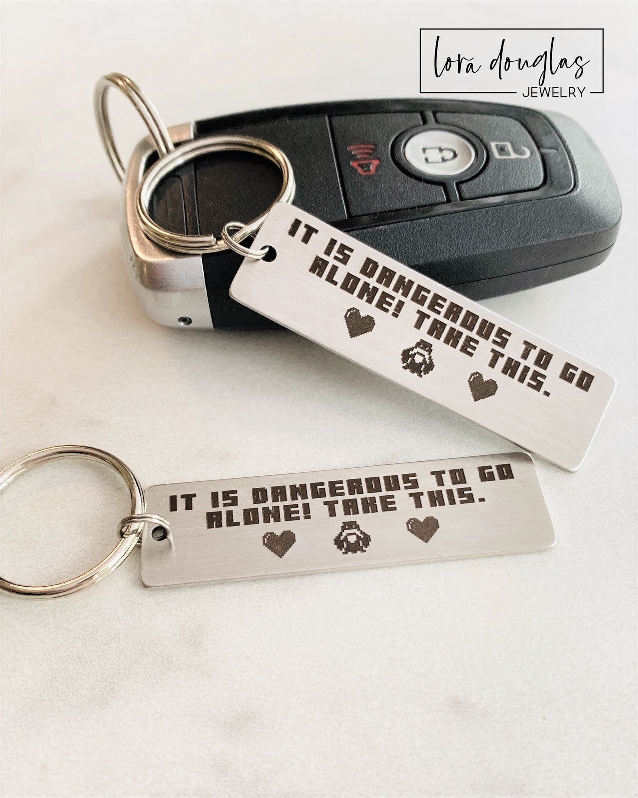 Gamer gift inspired by The Legend of Zelda featuring the famous "It is dangerous to go alone. Take this." quote engraved on a stainless steel keychain. Includes two pixel hearts and optional personalized engraving on the reverse for a unique keepsake.