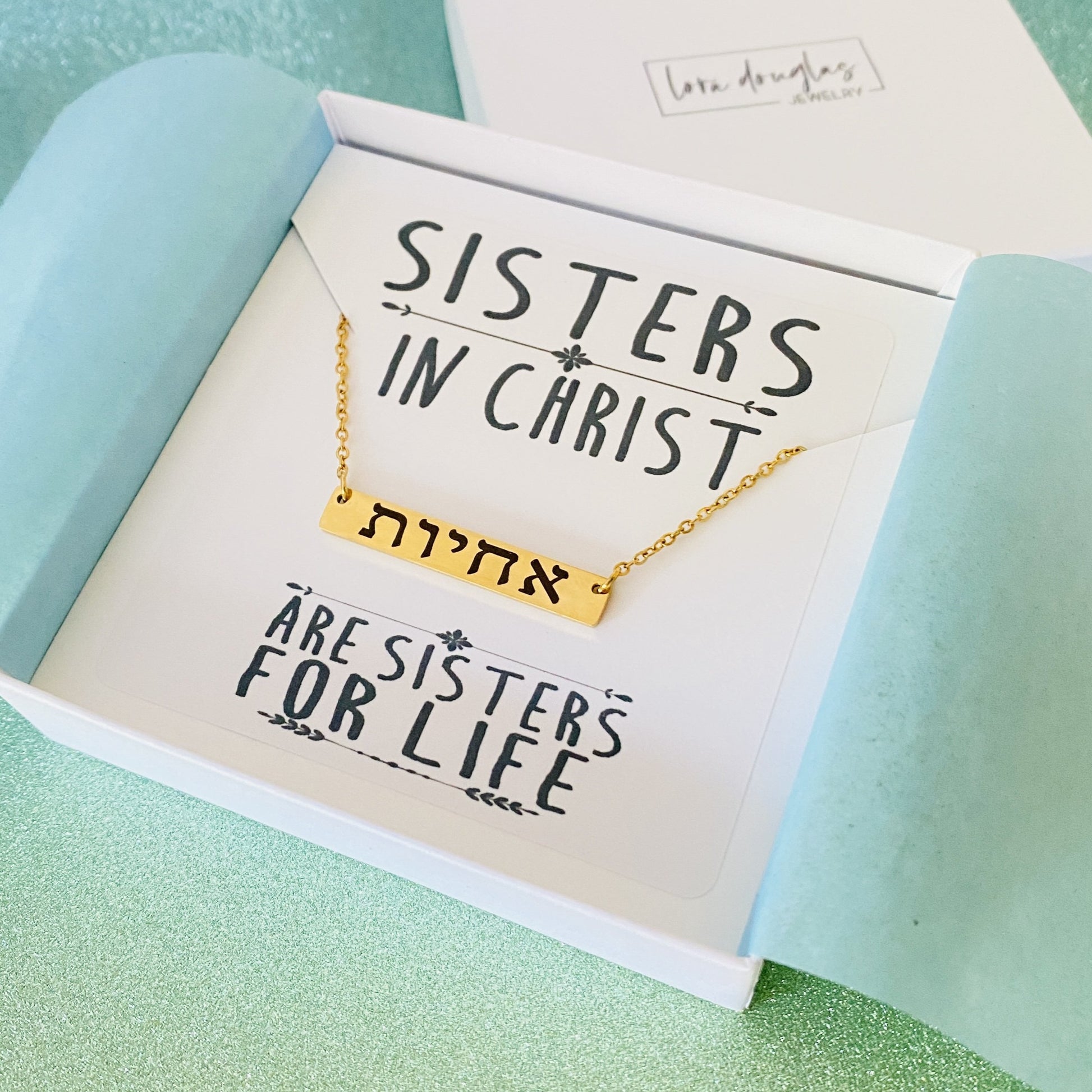 A gold bar necklace engraved with 'SISTERS IN CHRIST' in Hebrew script, presented in a box with the text 'SISTERS IN CHRIST ARE SISTERS FOR LIFE'. 