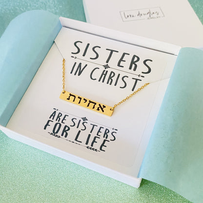 A gold bar necklace engraved with 'SISTERS IN CHRIST' in Hebrew script, presented in a box with the text 'SISTERS IN CHRIST ARE SISTERS FOR LIFE'. 