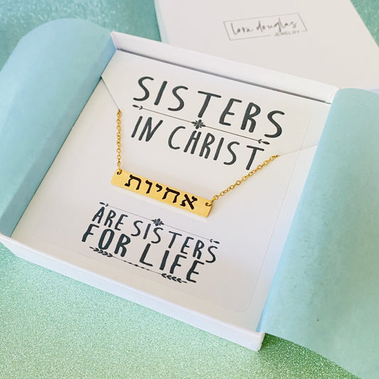 A gold bar necklace engraved with 'SISTERS IN CHRIST' in Hebrew script, presented in a box with the text 'SISTERS IN CHRIST ARE SISTERS FOR LIFE'. 