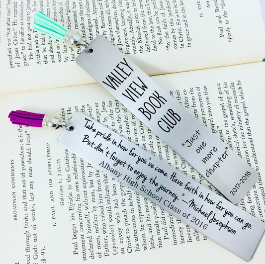 Two engraved metal bookmarks with personalized inscriptions, one in purple and one in teal, laying on an open book. Each bookmark has a tassel 