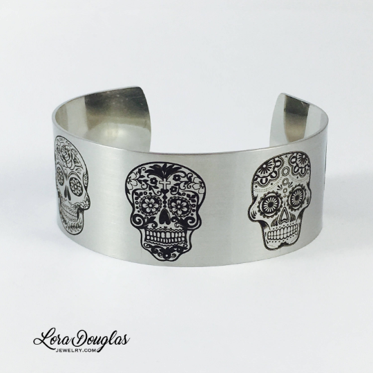 One inch wide sugar skull cuff bracelet made from stainless steel with five different engraved skull designs, available in silver and gold for collectors of Day of the Dead inspired, gothic, and alternative jewelry.
