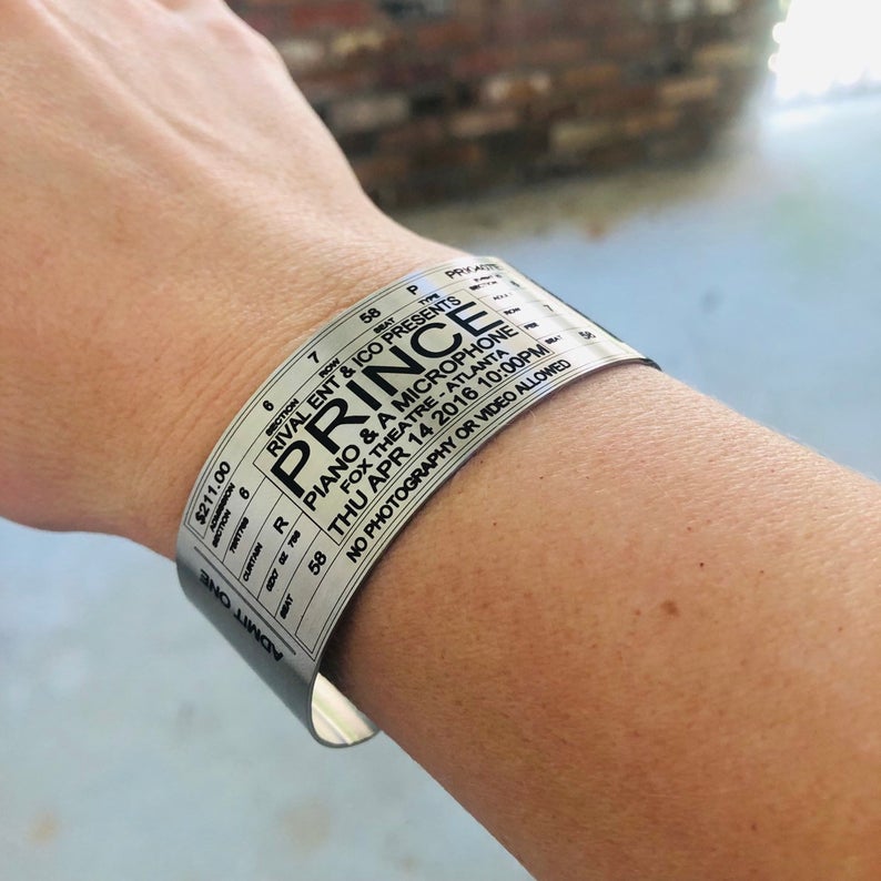 Personalized event ticket cuff bracelet showing custom artist, venue, date and seating information with dark engraving on brushed stainless steel, a unique concert memory gift for a music fan.
