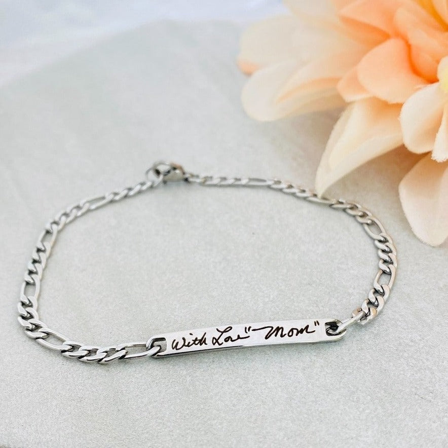 A stainless steel figaro chain bracelet with a plaque engraved with a handwritten message. 