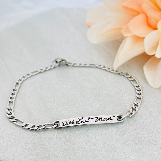 A stainless steel figaro chain bracelet with a plaque engraved with a handwritten message. 