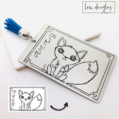 A custom stainless steel bookmark with a laser engraved child's drawing, presented next to its navy blue velvet drawstring pouch.